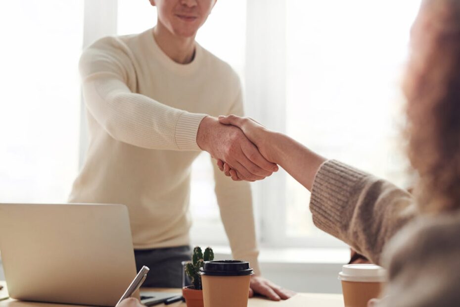 Two people in professional attire shaking hands over a modern desk where a real estate contract and house keys are laid out.