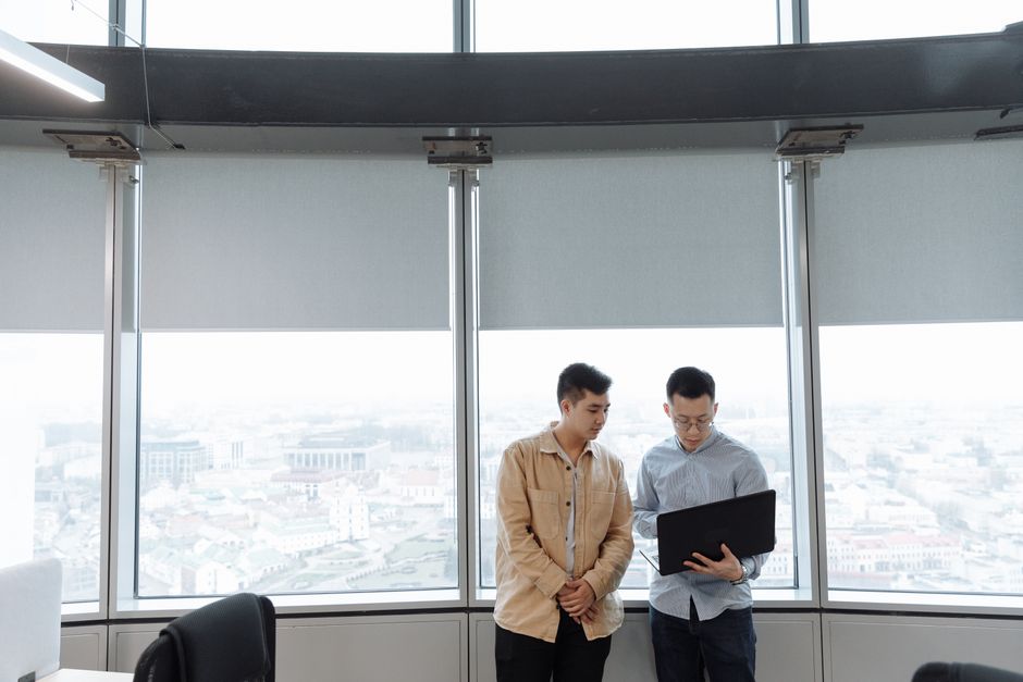 Well-dressed professional stands in a modern, high-rise office, looking thoughtfully out the window at a bustling city sky...